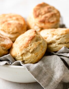 Tall and flaky biscuits stacked in a serving bowl with a grey kitchen cloth.