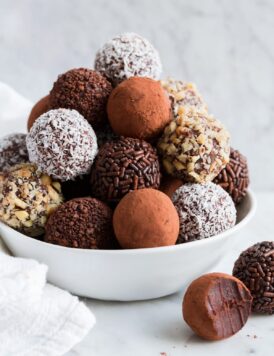 Stack of homemade chocolate truffles covered in cocoa, nuts, sprinkles and coconut piled into a pyramidal shape in a white bowl set over a marble surface.