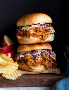 Photo: Two BBQ Pulled Chicken Sandwiches stacked on a wood platter. Apples slices and potato chips are shown to the side as serving suggestions.