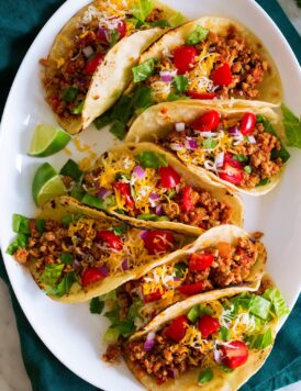 Photo: 7 ground turkey tacos in a row shown on a white oval platter from above. Tacos are filled with meat, tomatoes, lettuce, cheese and red onion.