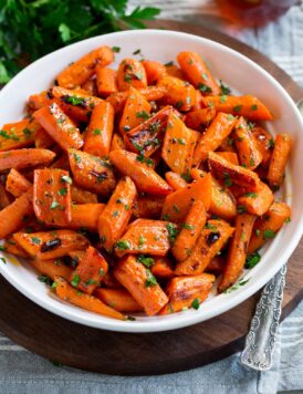 Photo: Roasted carrots in a white serving bowl. Carrots are garnished with parsley with a bunch in the background. Bowl is resting on a wooden platter and a grey cloth.