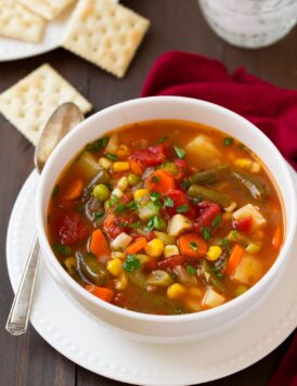 Single serving of vegetable soup in a white bowl set over a white plate. Set on a dark wooden table with a red napkin to the side.