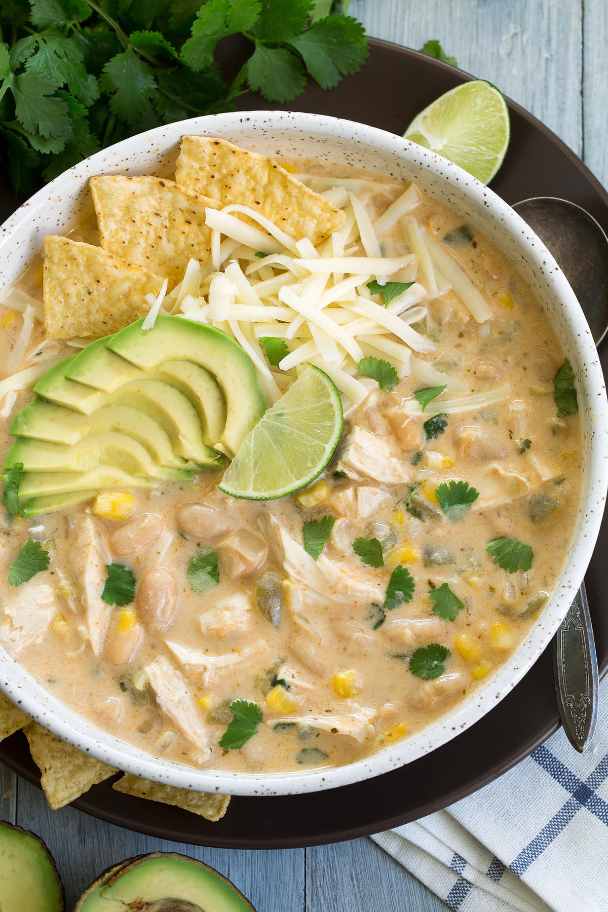 Overhead image of white chicken chili in a large white bowl sitting on a brown plate. Chili is garnished with cheese, chips, avocado, cilantro and lime.